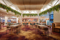 A spacious indoor lounge dining area with multiple high-top tables, bar stools, and warm wood ceiling panels. Artificial Green trailing plants hang from the ceiling, and the space is illuminated by bright overhead lighting.