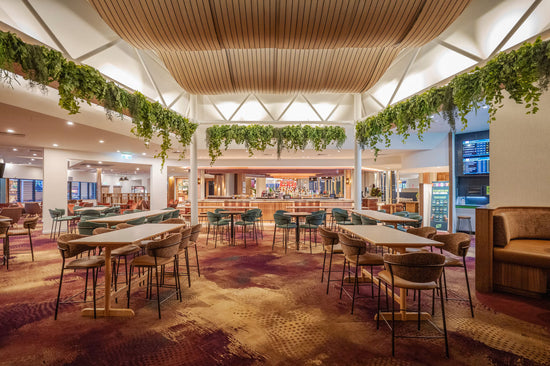 A spacious indoor lounge dining area with multiple high-top tables, bar stools, and warm wood ceiling panels. Artificial Green trailing plants hang from the ceiling, and the space is illuminated by bright overhead lighting.